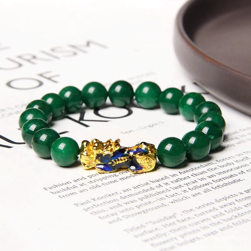 Pixiu jade protection bracelet displayed on dark ceramic tray with printed paper backdrop, showing forest green jade beads and gold Pixiu charm