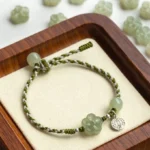 Handmade plum blossom jade prosperity bracelet with green braided rope, jade beads, and silver charm on wooden display tray