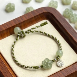 Handmade plum blossom jade prosperity bracelet with green braided rope, jade beads, and silver charm on wooden display tray