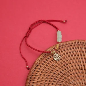 Red string jade Fu bracelet flat lay with adjustable braided ends and gold Fu charm on red background with rattan coaster