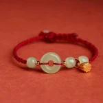 Red string jade peace buckle lotus bracelet - flat lay overhead view with light green jade donut, gold lotus charm, and jade beads on red background