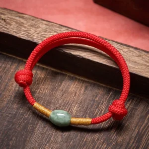 Red jade protection bracelet flat lay top-down view showing braided red cord with gold accents and oval green jade bead on wooden desktop