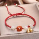 Red peace buckle jade lotus bracelet with red braided cord, gold lotus charm, and red agate beads on luxury satin background