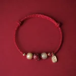 Red braided luck bracelet with Hetian jade beads and cinnabar lotus charm, 14K gold plated accents, flat lay on wine red background