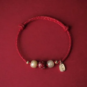 Red braided luck bracelet with Hetian jade beads and cinnabar lotus charm, 14K gold plated accents, flat lay on wine red background
