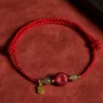 Red string cinnabar peace buckle jade bracelet with gold Fu charm displayed on red suede background, featuring natural jade beads