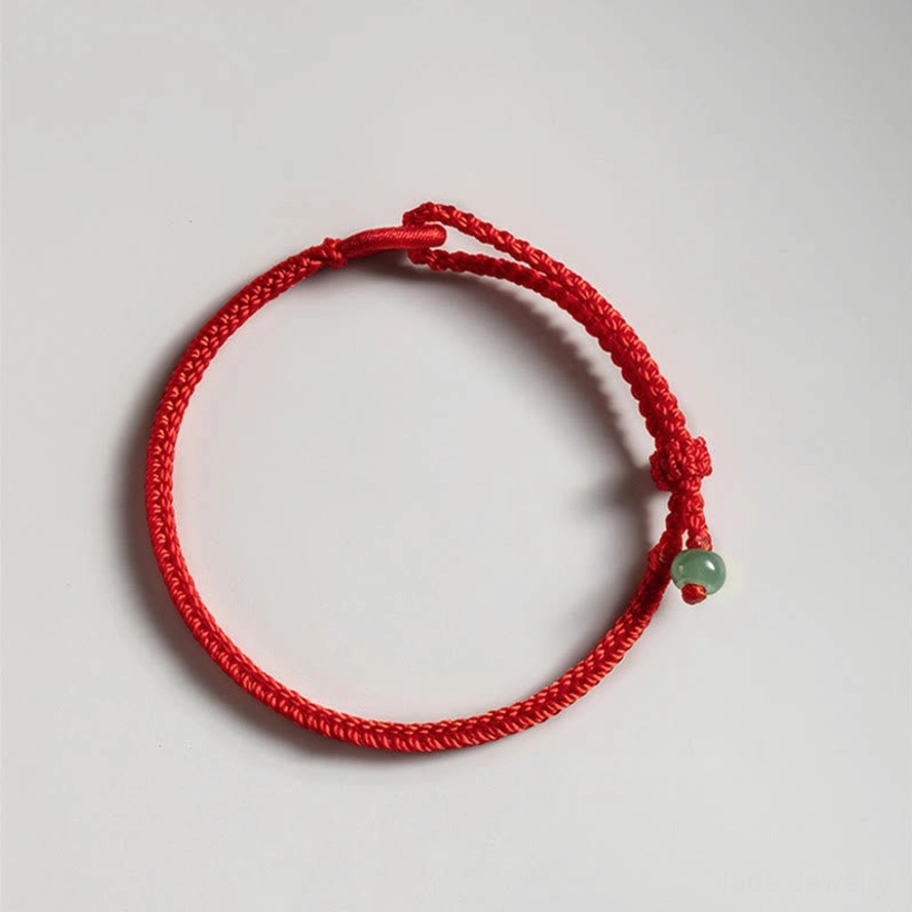 Single red string jade protection bracelet on light gray background, top-down view showing braided red rope, sliding adjustable knot, and small green jade pendant