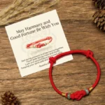 Red string jade bracelet with golden accents displayed on wooden surface with pinecones and matching blessing greeting card