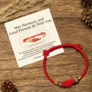Red string jade bracelet with golden accents displayed on wooden surface with pinecones and matching blessing greeting card