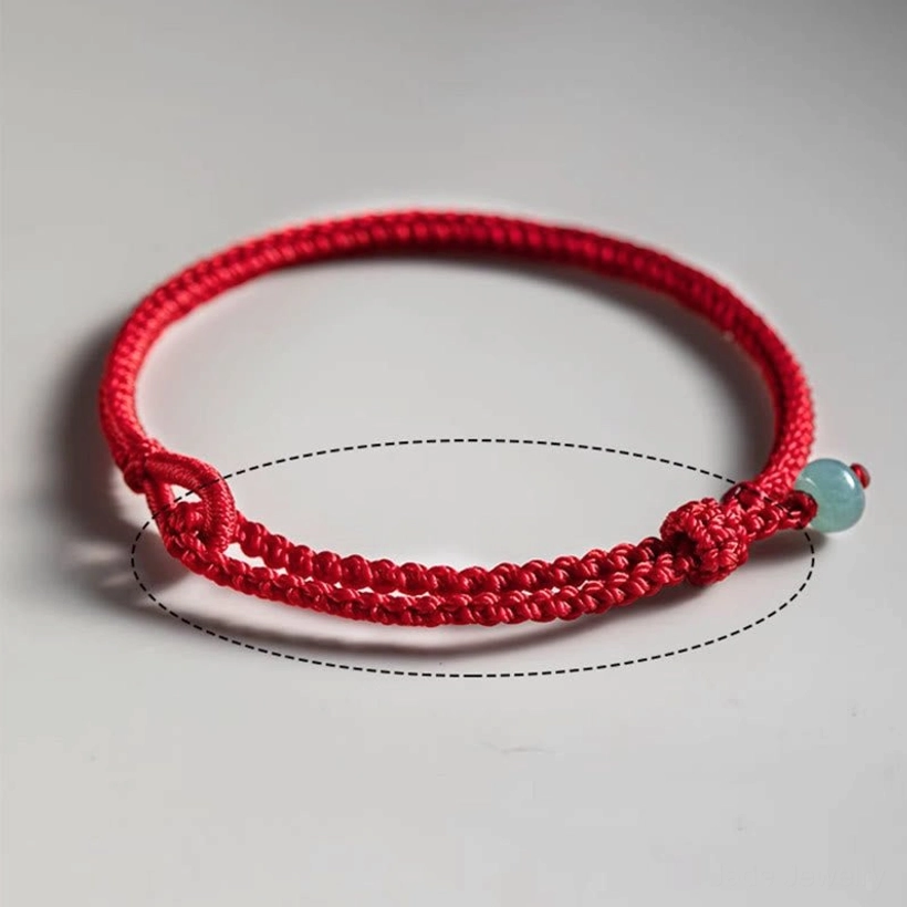 Red string jade bracelet overhead close-up on gray background, displaying clear braided texture, dashed sizing guide line, and light green jade charm detail