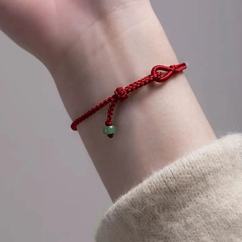 Red string jade bracelet on wrist with light knitted sweater background, showing prominent braided knot and jade pendant hanging at wrist