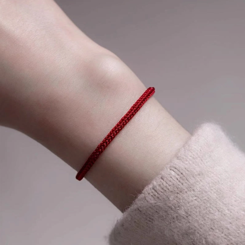 Red string jade bracelet worn on wrist with sweater cuff, close-up side view showing slim red cord and small hanging jade bead in snug fit