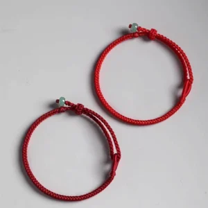 Two matching red string jade bracelets arranged on light gray background, displaying braided red ropes, green jade charms, and adjustable fastener knots