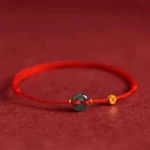 Red string jade peace buckle bracelet with copper coins on dark background, front flat view showing adjustable sizing