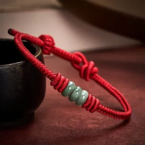 Two red jade protection bracelets with braided cord, jade bead clusters and decorative knots displayed on black ceramic cup