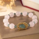 Rose quartz jade Pixiu beaded bracelet with gold filigree charm, flat lay on brown fabric with dried flowers