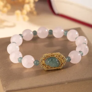 Rose quartz jade Pixiu beaded bracelet with gold filigree charm, flat lay on brown fabric with dried flowers
