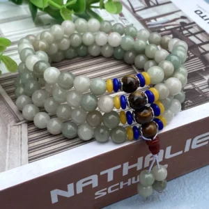 Top-view of 108 Shetai jade mala beads bracelet with light green natural jade beads, colorful accent spacers, and red tassel, displayed on open book with green foliage