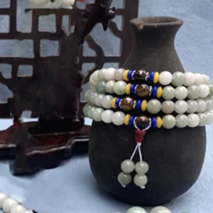 Close-up front detail of natural Shetai jade mala prayer beads bracelet showing smooth polished jade round beads with blue, yellow, and dark brown decorative accent spacers on ceramic vase