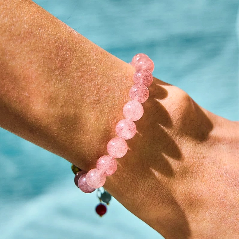 Pink strawberry quartz beaded bracelet with small red tassel charm on human wrist with water background