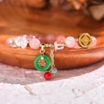 Strawberry quartz and jade fu blessing bracelet with pink crystal beads, green jade charm, and gold hardware on wooden surface