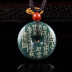 Dark teal jade peace buckle pendant with gold Taoist engravings and mountain ghost talisman, displayed on dark reflective surface with red accent bead and braided rope necklace