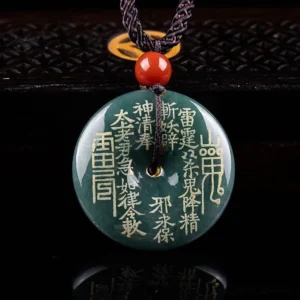 Dark teal jade peace buckle pendant with gold Taoist engravings and mountain ghost talisman, displayed on dark reflective surface with red accent bead and braided rope necklace