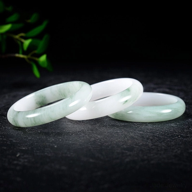 Three stacked natural jade bangles displaying varied green-white patterns and hand-polished surfaces with foliage background