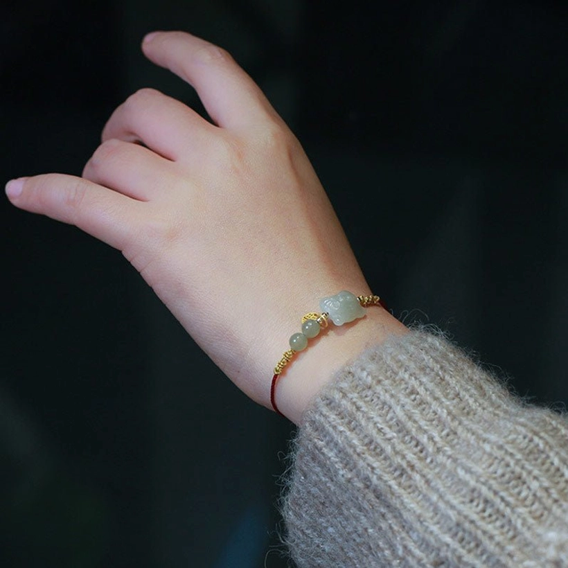 Adjustable zodiac tiger jade bracelet on finger displaying thin red cord, small jade beads, and delicate gold woven details