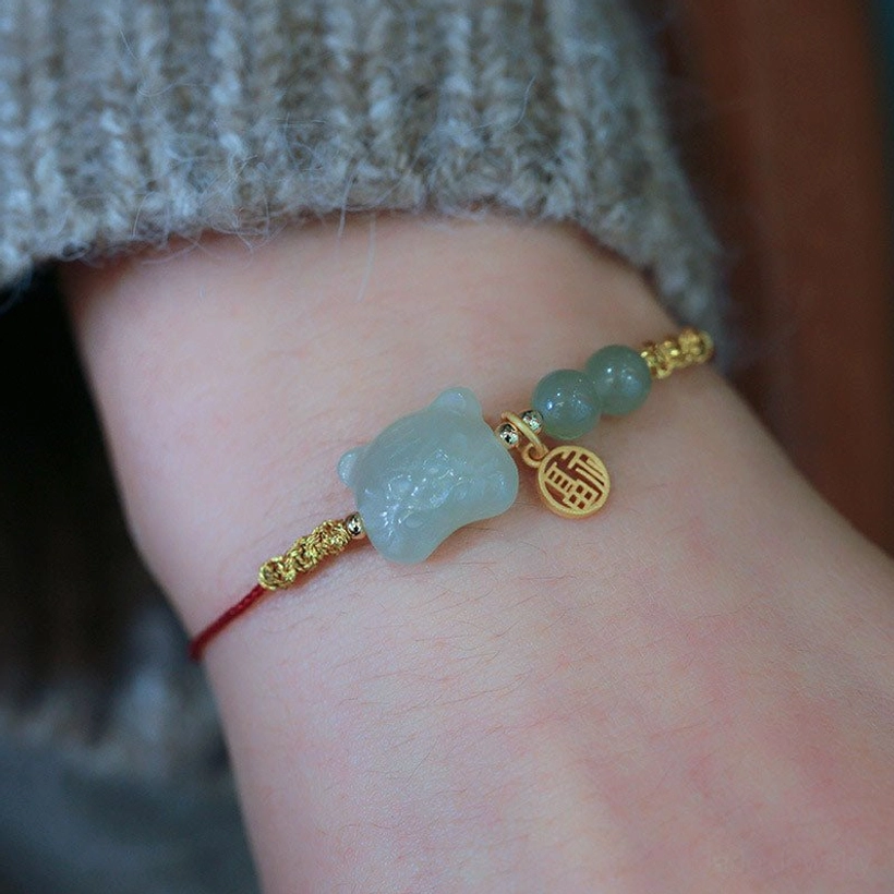 Tiger jade red string bracelet close-up top-down view on bare skin displaying oversized jade tiger bead, gold fortune tag, and multiple jade beads