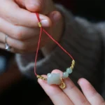 Tiger zodiac jade prosperity bracelet close-up displaying red woven cord, carved jade tiger charm, gold fortune pendant, and jade beads in hand