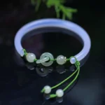 White Agate Hetian Jade interlocking bangle bracelet with green braided cord, overhead close-up on dark glossy surface with green plants