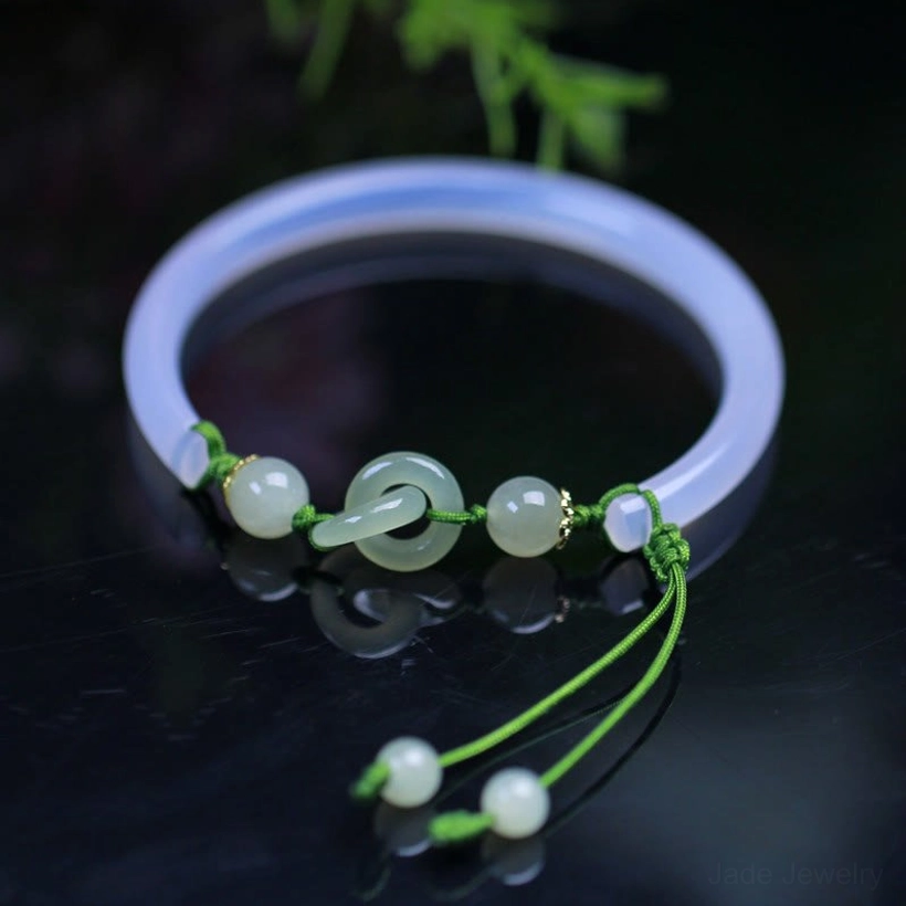 White Agate Hetian Jade interlocking bangle bracelet with green braided cord, overhead close-up on dark glossy surface with green plants