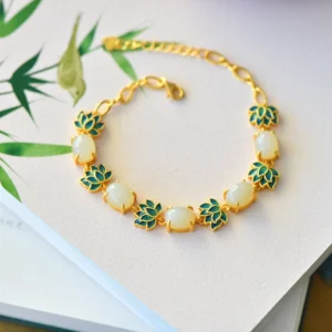 White jade and cyan jade lotus protection bracelet, top-down flat lay on book with bamboo leaves, showing white oval jade beads with blue enamel lotus charms and gold metal accents