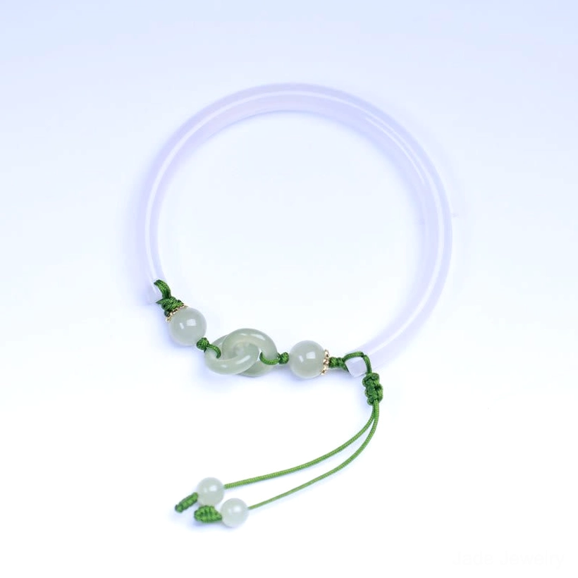 Single white Hetian jade interlocking bangle bracelet with green cord and dangling jade pendant, overhead flat lay on light blue background