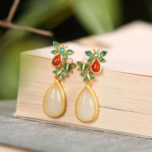 White jade butterfly teardrop earrings with red agate and enamel flower stud, displayed on wooden book background, showing gold-plated setting and pearl accents