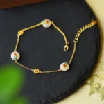 White jade protection bracelet with gold-plated copper chain displayed overhead on black slate, showing enamel-decorated jade beads and coin-shaped gold accents