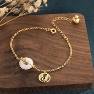 White jade double happiness bracelet flat lay with gold charm, showing adjustable chain and natural jade donut pendant on wooden coaster with dried flowers