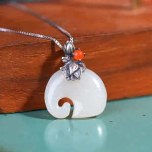 White jade elephant lotus necklace pendant displayed at oblique angle on wooden surface, showing polished jade carving with vintage silver lotus bail and red stone accent alongside silver box chain