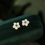 White jade flower stud earrings with green gem center in gold setting, displayed in dark green velvet jewelry case, top-down view