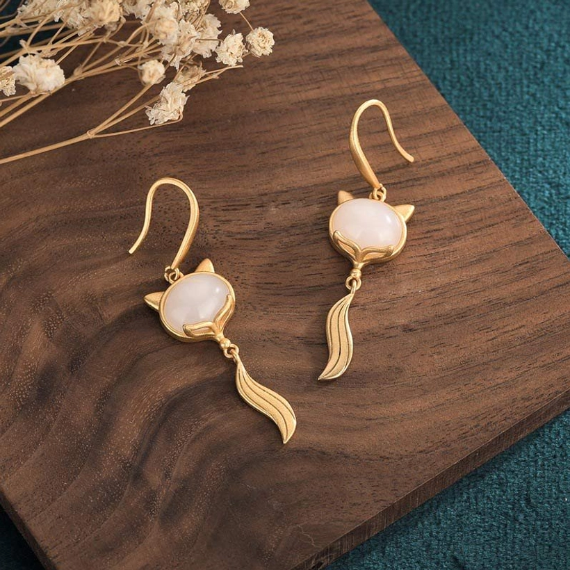 White jade fox earrings side view, separated pair on wooden plate with teal background, featuring gold ear hooks and flowing tail pendants