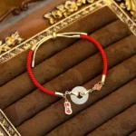 White jade fu character red string blessing bracelet with copper clasp displayed on gold-trimmed velvet tray, top-down side view