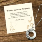 White jade koi fish necklace with twisted silver chain displayed on wooden surface next to personalized blessing message card, overhead flat lay view