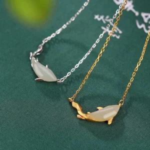 Pair of white jade koi fish necklaces in silver and gold variants displayed together, flat lay on dark green calligraphy background showing both style options