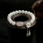 Natural white jade luck bracelet with shell flower charm and dual teardrop pendants, top-down view on dark wooden display holder