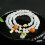 White jade peace buckle Fu character double-layer bracelet with golden silk jade accents and orange beads displayed on dark wooden tray with green foliage