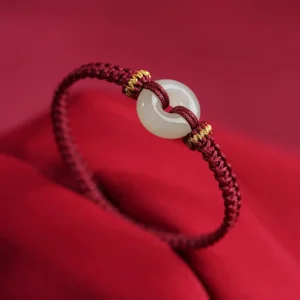 White jade peace buckle donut pendant on wine-red braided cord bracelet with gold knot accents, three-quarter view on velvet