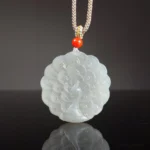 White jade peacock pendant necklace flat lay overhead view showing intricate carved peacock feather design on light grey jade with braided cord and red protective bead