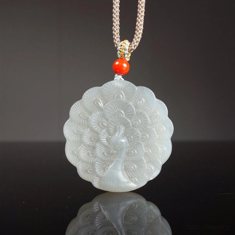 White jade peacock pendant necklace flat lay overhead view showing intricate carved peacock feather design on light grey jade with braided cord and red protective bead