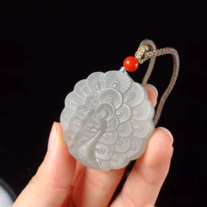 White jade peacock necklace held in hand showing size and delicate braided cord with red accent bead against dark background
